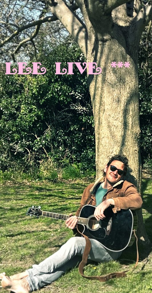 Image of Lee Probert with a guitar sitting on grass under a tree